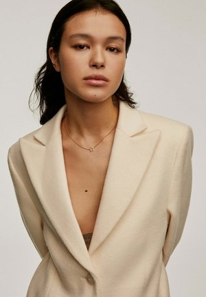 Blazer de lana beige con escote en V profundo, cierre de un solo botón y solapas afiladas, complementado con un delicado collar de estrella dorada.
