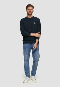 Donkerblauwe sweatshirt met ronde hals en ribgebreide manchetten, gecombineerd met lichtblauwe jeans en witte sneakers, staand tegen een neutrale achtergrond.