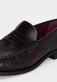 Μαύρο δερμάτινο penny loafer με ραφές μοκασίνου, στρογγυλεμένη μύτη, χαμηλό τακούνι και μπορντό εσωτερική επένδυση.