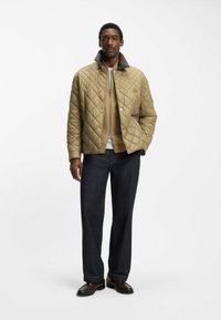 Giacca trapuntata color cachi con colletto marrone, cerniera frontale e bottoni a pressione, indossata sopra un maglione beige e jeans scuri. Scarpe marroni completano il look.