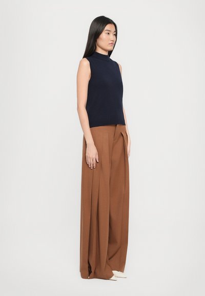 Jūras spēku zils bez piedurknēm turtlenecks tops, kas apvienots ar brūnām platajām biksēm ar krokām un gludu tekstūru, pabeigts ar gaišu krāsu apaviem.