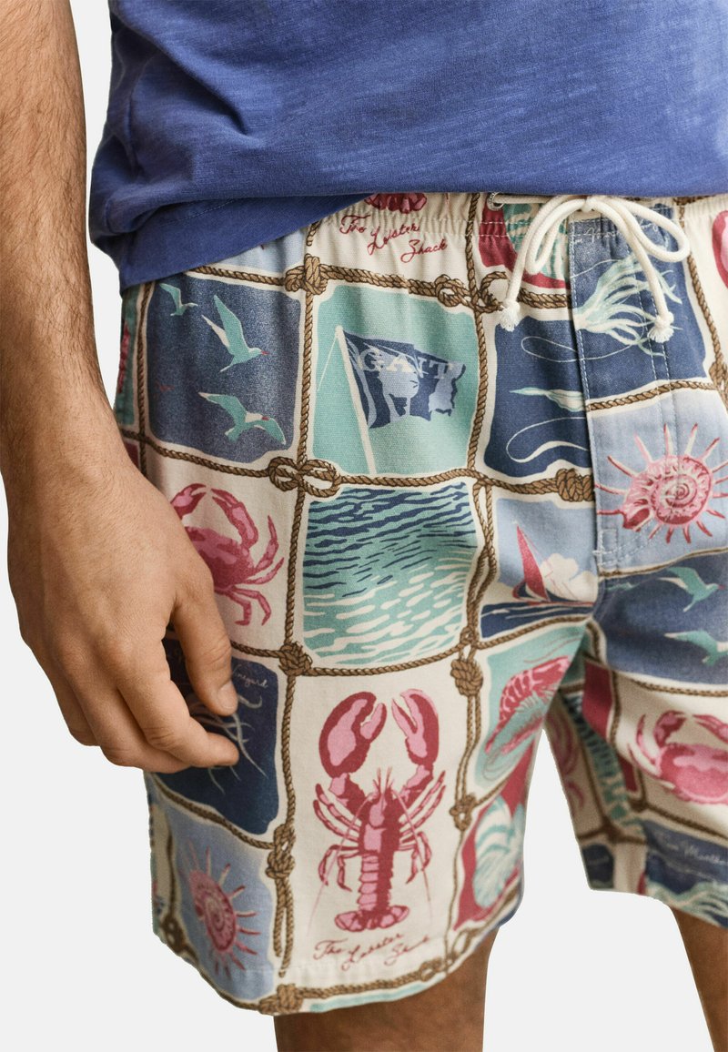 Homme portant un short à thème nautique avec un imprimé de homards, crabes, mouettes, coquillages et scènes océaniques, associé à une chemise bleue.