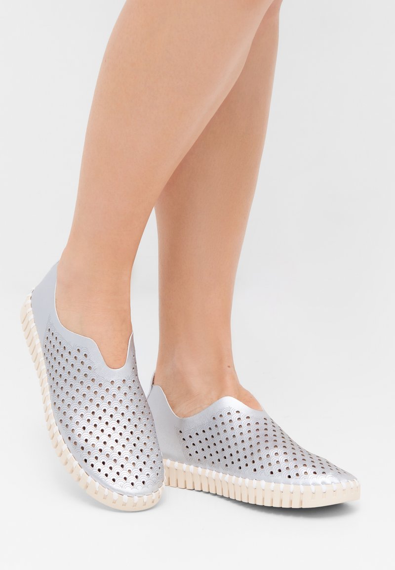 Zapatos plateados perforados tipo slip-on con laterales elásticos, suela de goma blanca y diseño de punta redondeada con un patrón texturizado.