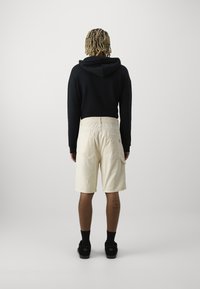 Personne aux cheveux blonds torsadés portant un sweat à capuche noir, un short beige aux genoux, des chaussettes noires et des chaussures noires, debout devant un fond uni.