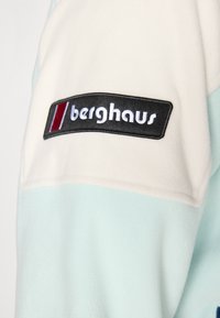 Primer plano de la manga de una chaqueta de forro polar blanca y azul claro con un parche negro del logo de Berghaus cosido.