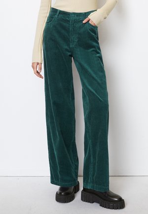 Kvinde iført mørkegrønne vidde-buks i fløjl med sorte chunky støvler, stående mod en hvid væg, hånd i lommen.