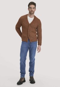 Brauner, gestrickter Cardigan mit V-Ausschnitt und Knopfverschluss, kombiniert mit blauen Jeans und schwarzen Schnürstiefeln. Darunter ein schlichtes weißes T-Shirt.