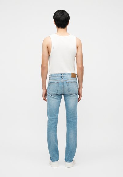 Άτομο που στέκεται με την πλάτη γυρισμένη, φοράει λευκό αμάνικο πουκάμισο, ανοιχτόχρωμο μπλε τζιν και λευκά αθλητικά σε απλό λευκό φόντο.