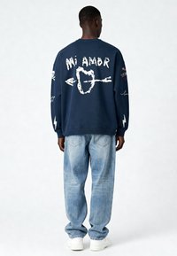 Sweatshirt bleu marine avec le texte graphique blanc "Mi Amor" et des skulls. Porté avec un jean bleu clair et des baskets blanches.