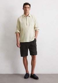 Linnen, lichtgele overhemd met lange mouwen, een knoopsluiting en een zak; gecombineerd met donkere shorts met cargozakken; zwarte instappers.
