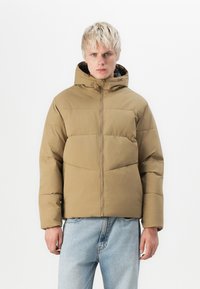Khaki puffer jacket with a zipper front, hood, and quilted design; paired with light blue denim jeans. Smooth texture; casual fit.