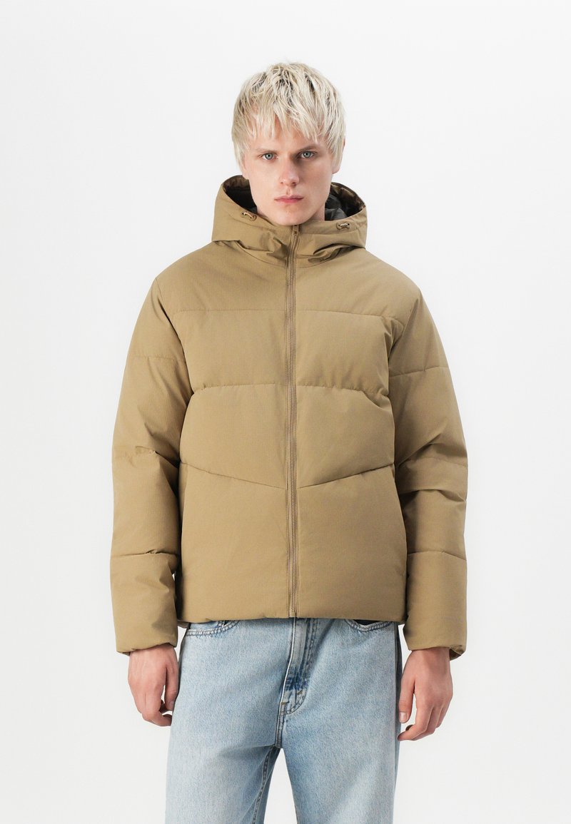 Khaki puffer jacket with a zipper front, hood, and quilted design; paired with light blue denim jeans. Smooth texture; casual fit.