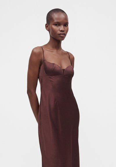 Femme aux cheveux courts portant une robe en satin marron foncé sans manches avec de fines bretelles, se tenant devant un fond clair uni.