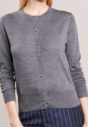 Femme portant un cardigan gris ajusté à boutons avec manches longues et un pantalon bleu marine à fines rayures.