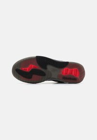 Semelle de chaussure Reebok présentant un design en caoutchouc texturé de couleur noire, avec des accents rouges et des motifs de semelle complexes pour une adhérence améliorée.
