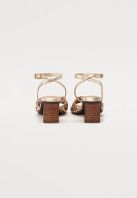 Sandales métalliques dorées avec un talon bloc, présentant un design tissé au niveau des orteils et des courroies à la cheville croisées à l'arrière. Talon texturé marron.