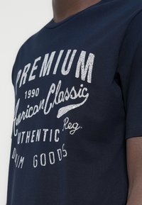 Μπλε βαμβακερό T-shirt ναυτικού στιλ με λευκό, υφασμένο κείμενο και γραφικά εκτυπωμένα με οθόνη, με στρογγυλή λαιμόκοψη και κοντά μανίκια.