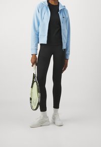 Sudadera azul claro con cremallera, camiseta negra, leggings negros, zapatillas blancas y sosteniendo una raqueta de tenis con un acento amarillo.