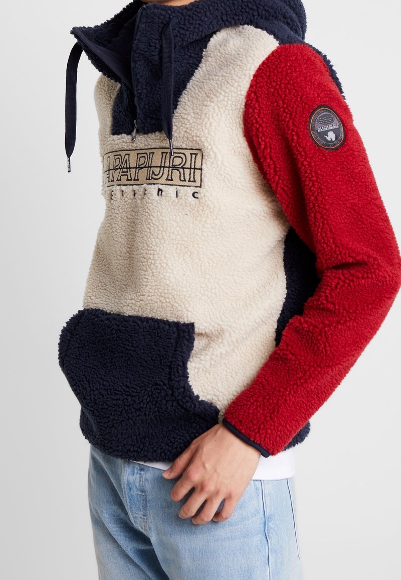Homme portant un sweat à capuche en polaire color block bleu marine, crème et rouge, avec une poche kangourou à l'avant et un logo brodé sur la poitrine et la manche.