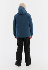Dunkelblaue isolierte Jacke mit Kapuze, elastischen Bündchen und lockerer Passform, getragen über einer schwarzen Hose. Eine beige Strickmütze ist sichtbar auf dem Kopf.