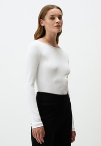 Weißes geripptes Langarmshirt mit weitem Ausschnitt, kombiniert mit schwarzen hochgeschnittenen Hosen. Glatte Textur, schmale Passform und minimalistisches Design.