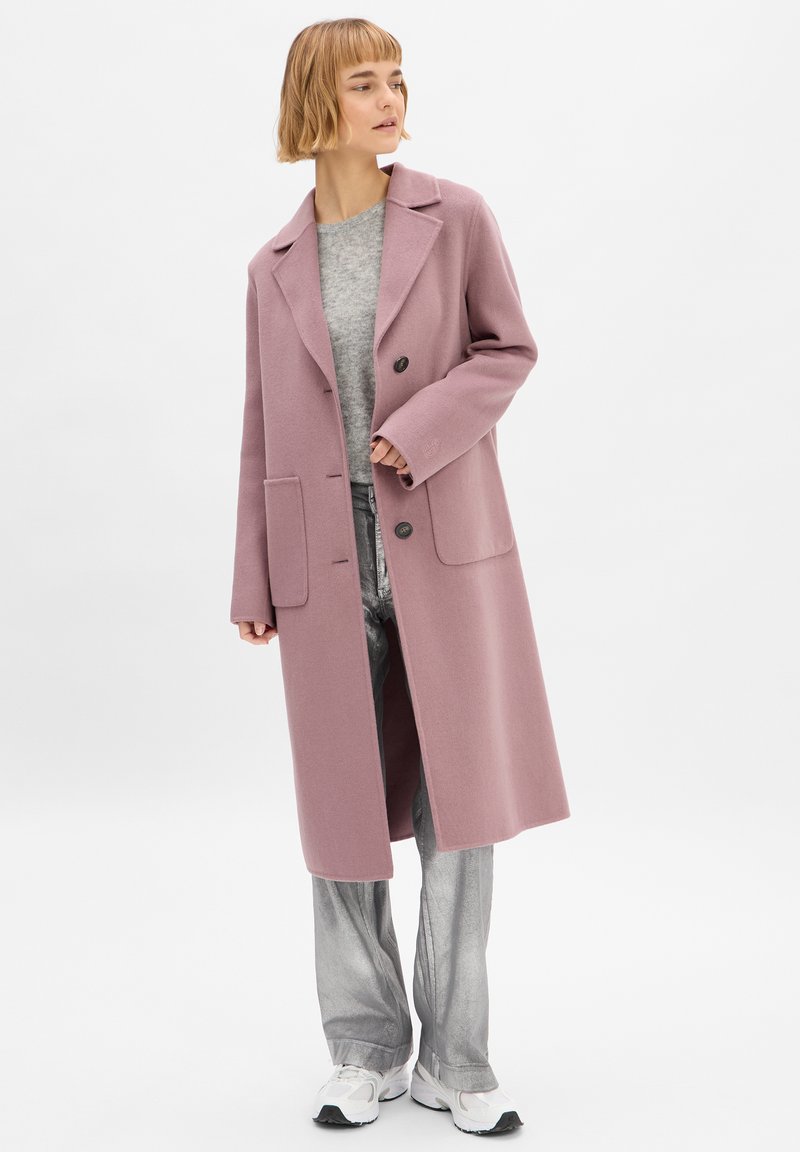 Cappotto di lana rosa chiaro con colletto a vista, due tasche frontali e chiusura con bottoni. Abbinato a una maglietta grigia e pantaloni larghi argentati.