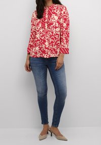 Blusa a fiori rossi con maniche lunghe e dettagli plissettati davanti, abbinata a jeans skinny blu e scarpe con tacco beige.