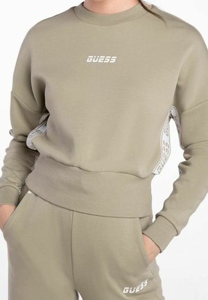 Beżowa bluza Guess i dopasowane spodnie dresowe z prążkowanymi mankietami, paskiem w pasie oraz logo na piersi i lewej udzie, noszone z rękami w kieszeniach.