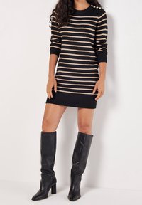 Femme portant une robe-pull à rayures noires et beiges avec des boutons dorés sur l'épaule et des bottes en cuir noires à talons montant jusqu'aux genoux.