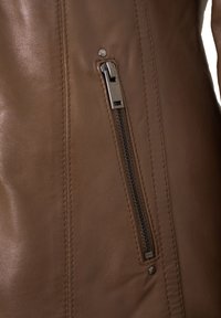 Nahaufnahme einer braunen Lederjacke mit seitlichem Reißverschluss, glatter Textur, sichtbaren Nähten und einem metallischen Reißverschlussgriff.