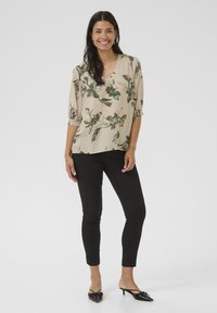 Top beige con motivo floreale verde, scollatura a V e maniche 3/4. Abbinato a pantaloni neri aderenti e ballerine nere a punta.