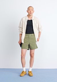 Ελαφρύ, ριγέ πουκάμισο μακρύ μανίκι πάνω από ένα μαύρο μπλουζάκι; πράσινα σορτς; ανοιχτόχρωμα, υφασμένα παπούτσια με καουτσούκ σόλες; λευκές κάλτσες.