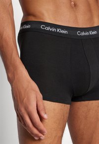 Schwarze Baumwoll-Boxershorts mit einem breiten elastischen Bund, auf dem "Calvin Klein" in weißen Buchstaben steht. Glatte Textur und eng anliegendes Design.