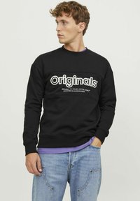 Sudadera negra con el texto blanco "originales" en el pecho, con cuello redondo y puños acanalados, llevada sobre una camisa morada.