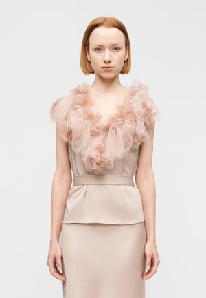 Młoda kobieta o prostych rudych włosach nosi sukienkę z różowego satynu z warstwowymi tiulowymi kwiatowymi zdobieniami wokół dekoltu i krótkich rękawach.