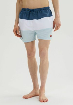 Hombre con bañador tipo color block en azul, blanco y azul claro con cordón, de pie descalzo sobre un fondo blanco.