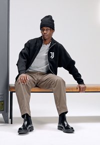 Zwarte jas met wit logo, grijs T-shirt, beige broek, zwarte leren laarzen. Zittend op een bankje in een minimalistische omgeving.