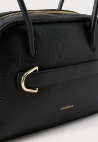 Bolso de mano de cuero negro texturizado con un adorno metálico dorado, asas dobles y cierre de cremallera visible con el logotipo grabado en el lateral.