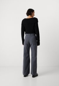 Rövid göndör hajjal rendelkező nő fekete vállra eső pulóverben, szürke, bő szárú nadrágban, fekete cipőben, egy sima fehér háttér előtt áll.