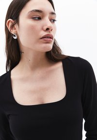 Schwarzes langärmliges Top mit quadratischem Ausschnitt. Der Stoff wirkt glatt und eng anliegend. Silberschmuck im Ohr des Modells ergänzt das Design.