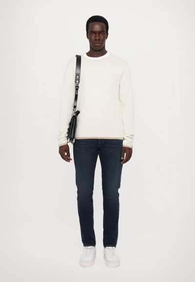 Weißer gerippter Pullover, dunkelblaue Jeans und weiße Sneaker. Schwarze Tasche mit Tragegurt. Model posiert vor einem hellgrauen Hintergrund.
