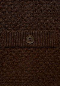 Material tricotat texturat de culoare maro, cu un tiv zimțat și un nasture maro închis cu patru găuri, creând un aspect stratificat și tactil.
