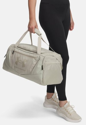 Beige duffeltas met dubbele handvatten, logo aan de voorkant, ritssluitingvakken en gestructureerde stof, vastgehouden door een persoon in zwarte leggings.