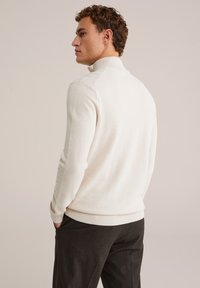 Muška off-white pletena dukserica s visokim ovratnikom, rebrastim manžetama i donjim rubom. Tekstura tkanine s suptilnim uzorkom, u kombinaciji s tamnim hlačama.