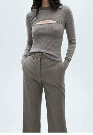 Kobieta w taupe prążkowanym swetrze z wycięciem na klatce piersiowej oraz brązowych, szerokich spodniach w prążki z wysokim stanem, ręce w kieszeniach, neutralne tło.