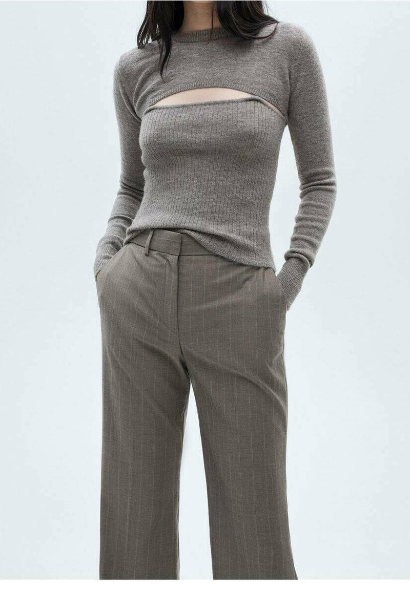 Femme portant un pull en maille côtelée taupe avec découpe poitrine et pantalon large à taille haute marron à fines rayures, mains dans les poches, fond neutre.