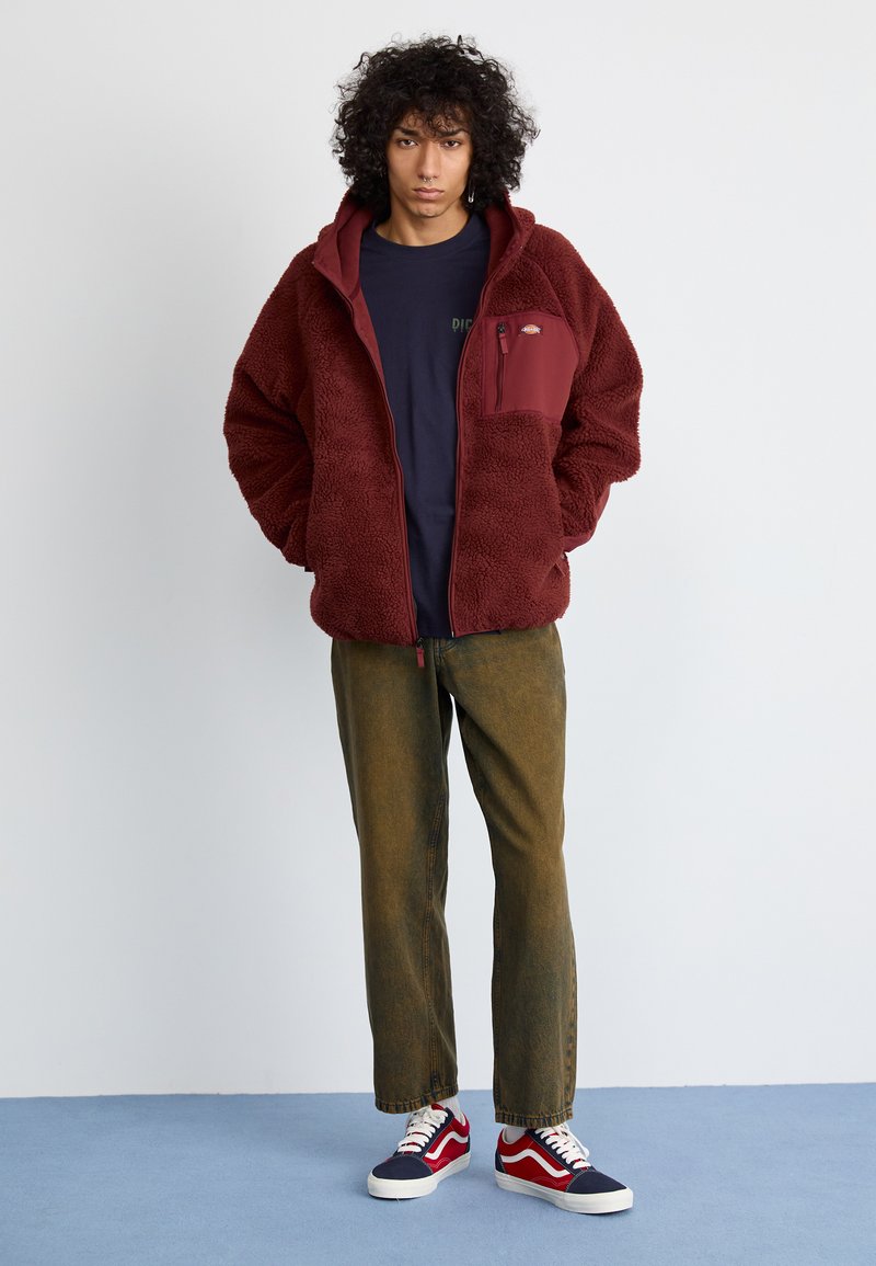 Veste en polaire bordeaux avec une capuche et une fermeture éclair à l'avant, comportant une poche poitrine. Portée sur une chemise bleu foncé et associée à un pantalon vert olive et des baskets.