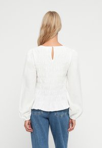 Hvid bluse med smocket ryg, rund halsudskæring og nøglehul detalje. Lange puffærmer, blød tekstur og flæsekant. Sammen med bukser i blå denim.