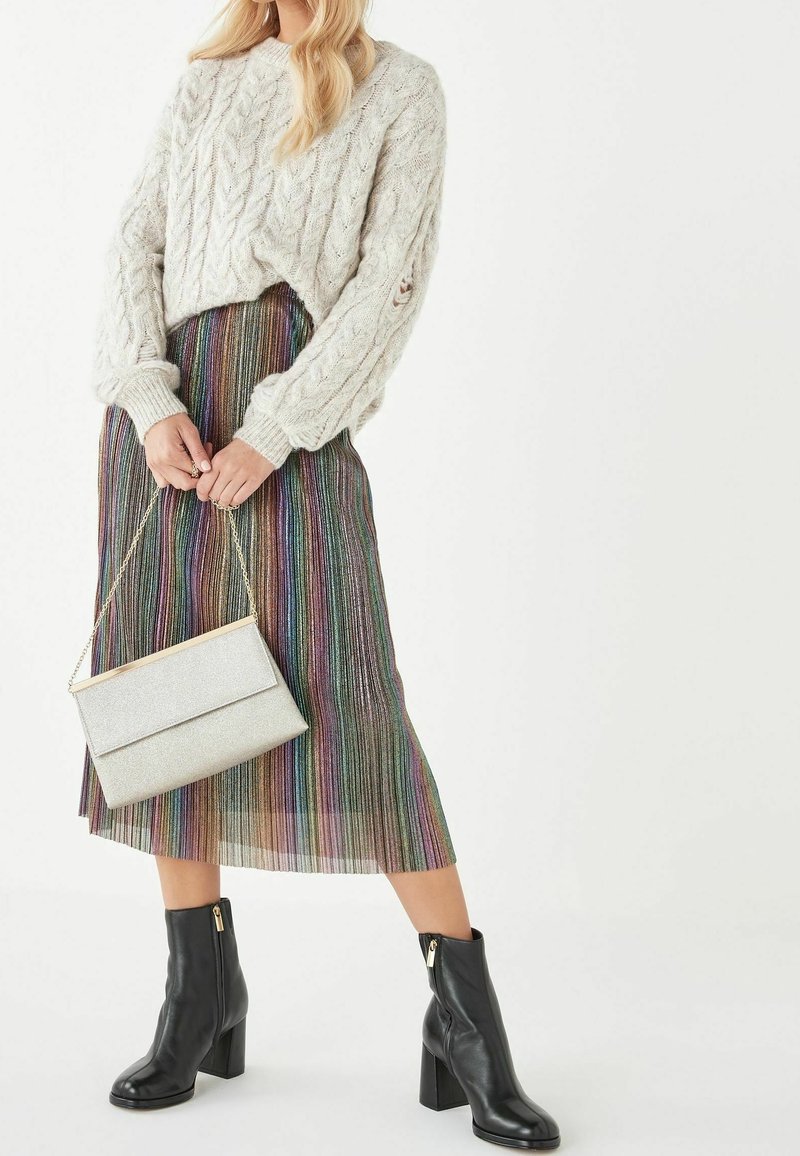 Personne portant un pull épais gris à torsades, une jupe midi plissée multicolore, des bottines noires à talons et tenant un sac à chaîne argenté.