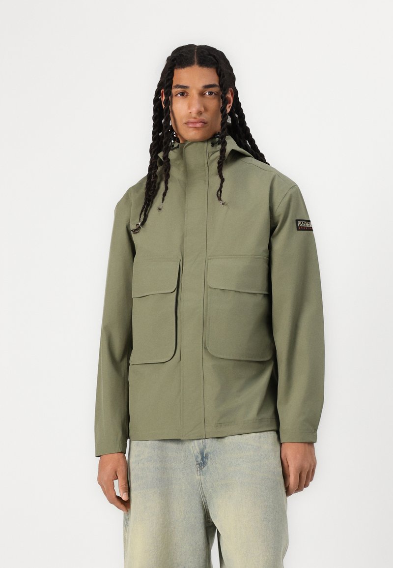 Chaqueta verde oliva con cuello alto, dos bolsillos delanteros, textura suave y capucha ajustable. Presenta un parche de logo negro en la manga.